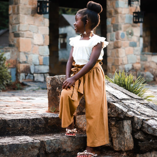 A young girl wearing a beige wide-legged paper sack pants set with a linen shirt and ruffle sleeves, sitting outdoors.